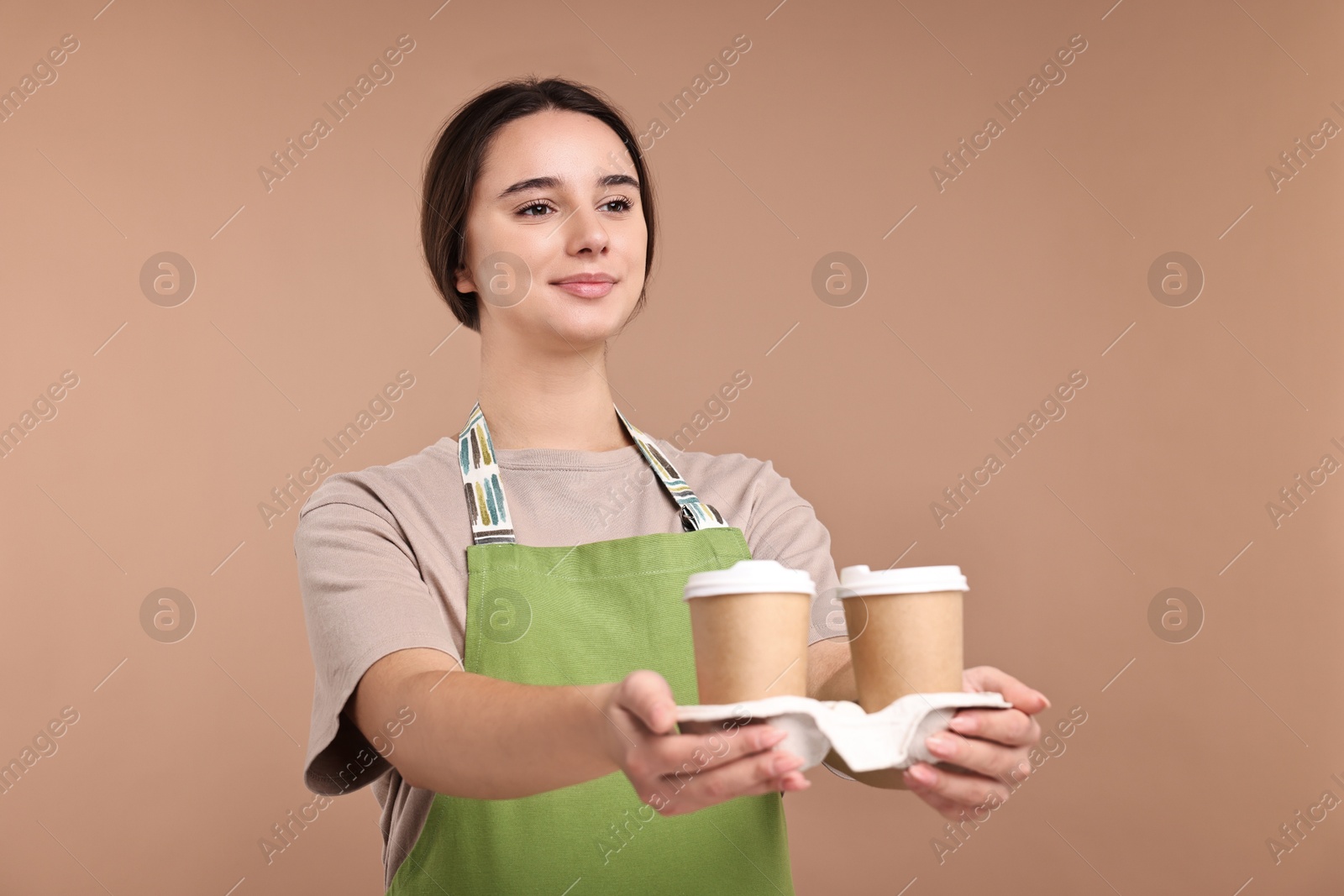 Photo of Girl in apron with takeaway paper cups of coffee on pale brown background. Work for teenagers