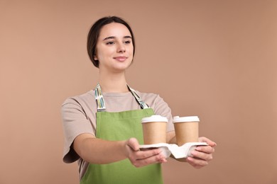 Photo of Girl in apron with takeaway paper cups of coffee on pale brown background. Work for teenagers