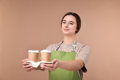 Photo of Girl in apron with takeaway paper cups of coffee on pale brown background. Work for teenagers