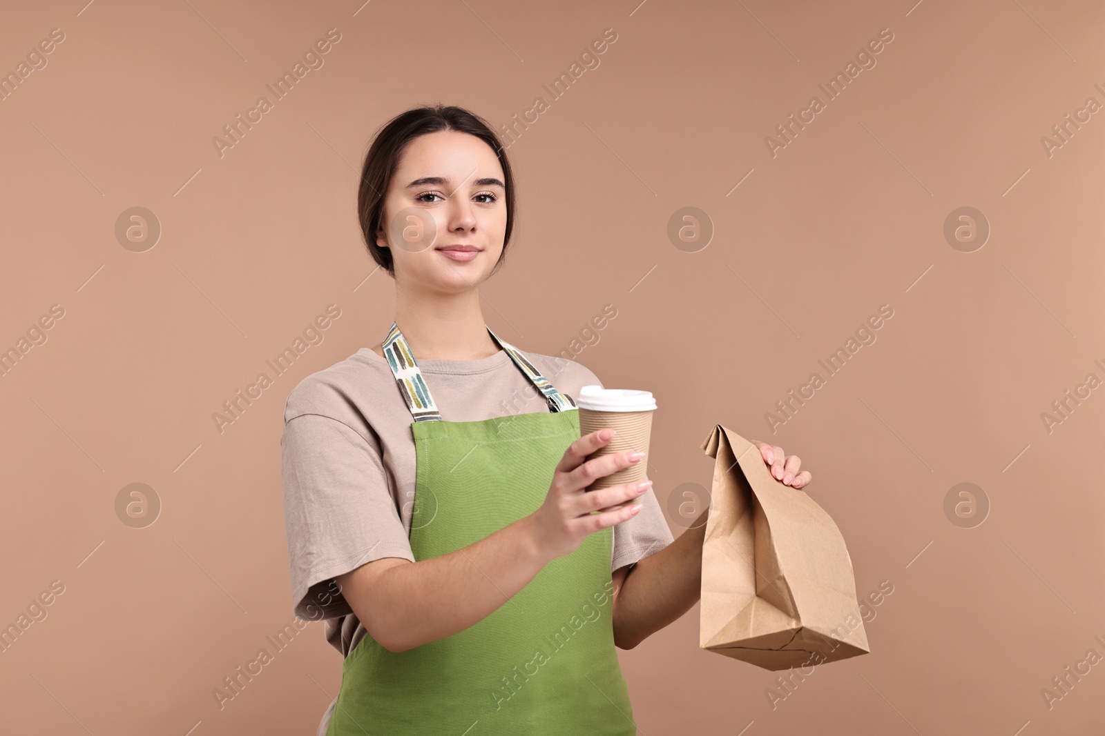 Photo of Girl in apron holding takeaway cup of coffee and paper bag on pale brown background. Work for teenagers