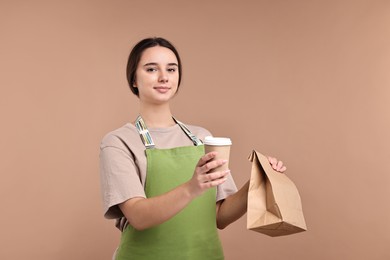 Photo of Girl in apron holding takeaway cup of coffee and paper bag on pale brown background. Work for teenagers