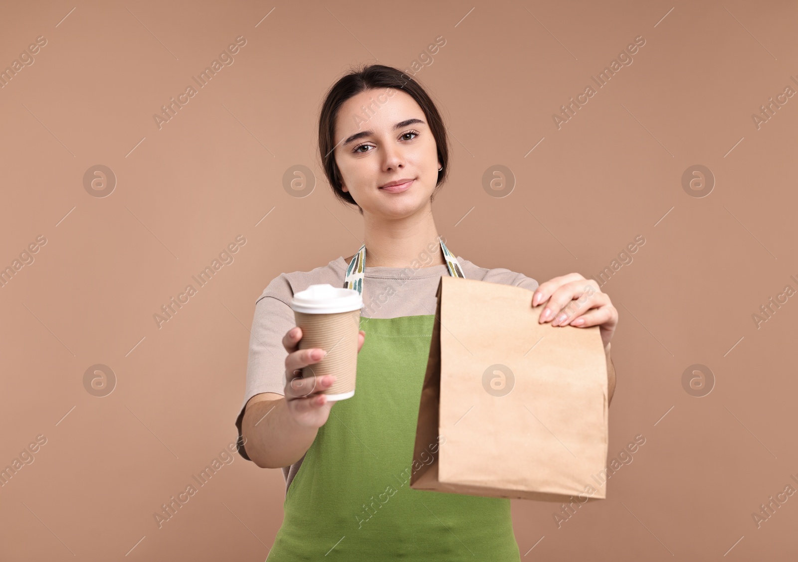 Photo of Girl in apron holding takeaway cup of coffee and paper bag on pale brown background. Work for teenagers