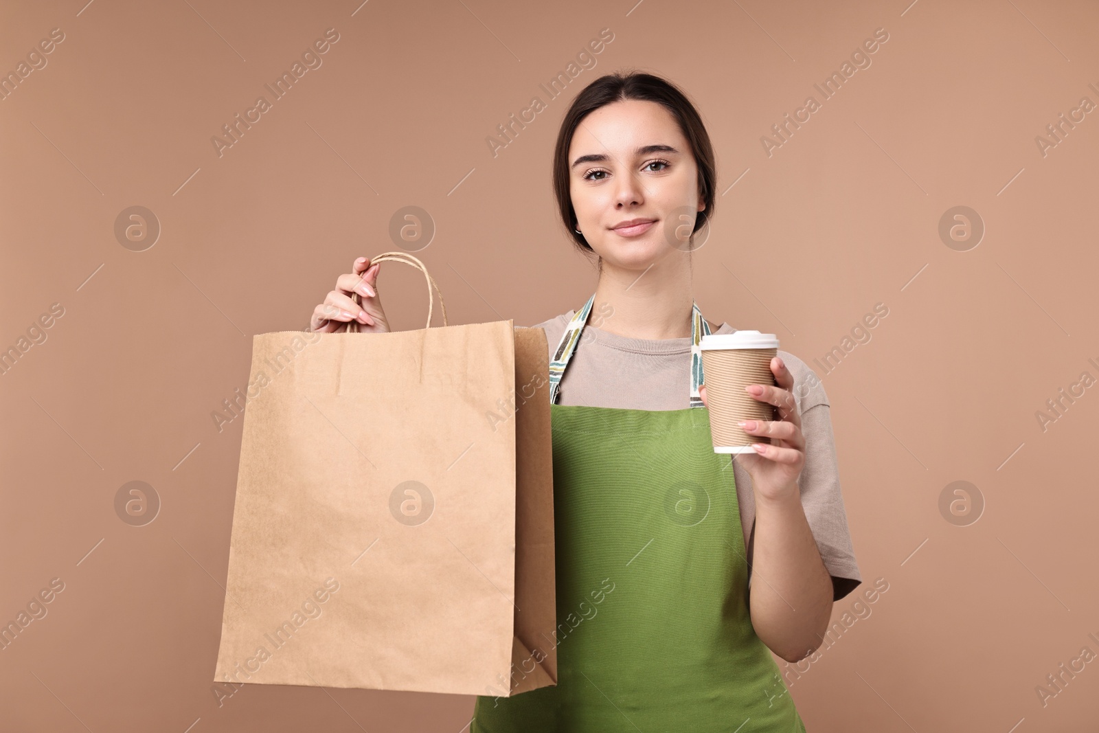 Photo of Girl in apron holding takeaway cup of coffee and paper bag on pale brown background. Work for teenagers