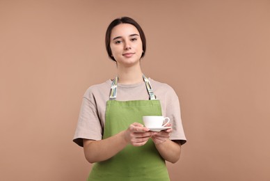 Photo of Girl in apron with cup of coffee on pale brown background. Work for teenagers