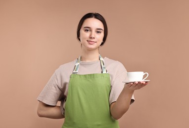 Photo of Girl in apron with cup of coffee on pale brown background. Work for teenagers