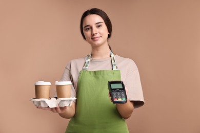 Photo of Girl in apron with takeaway paper cups of coffee and payment terminal on pale brown background. Work for teenagers