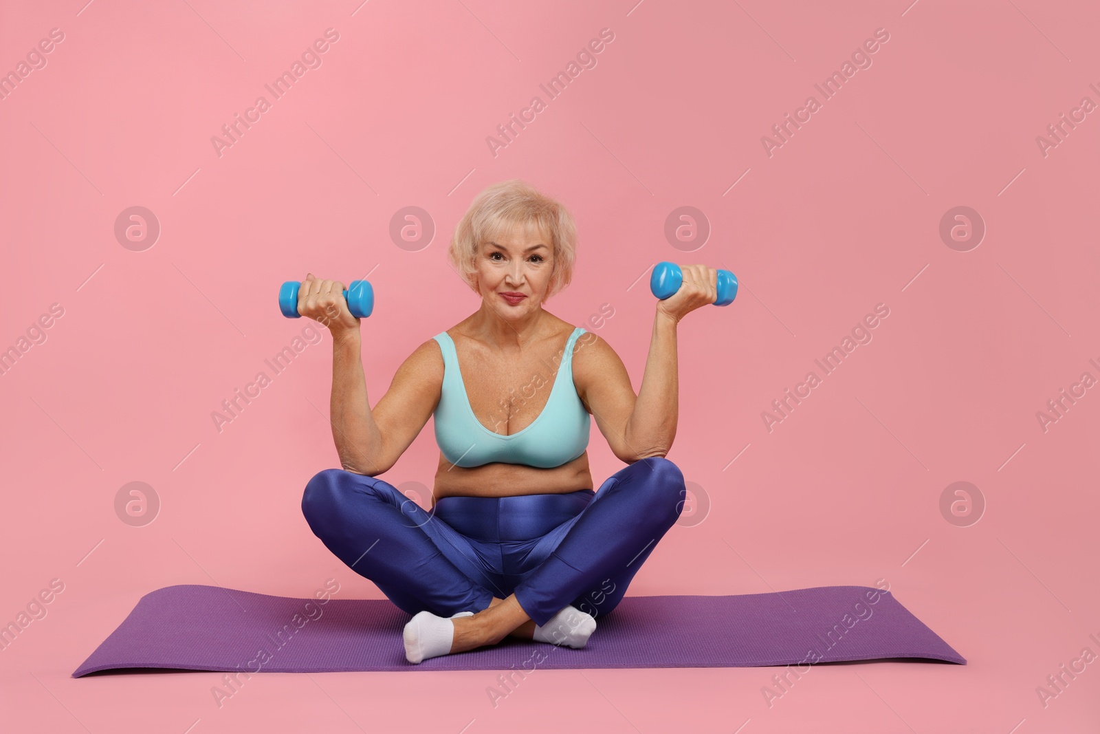 Photo of Senior woman exercising with fitness mat and dumbbells on pink background