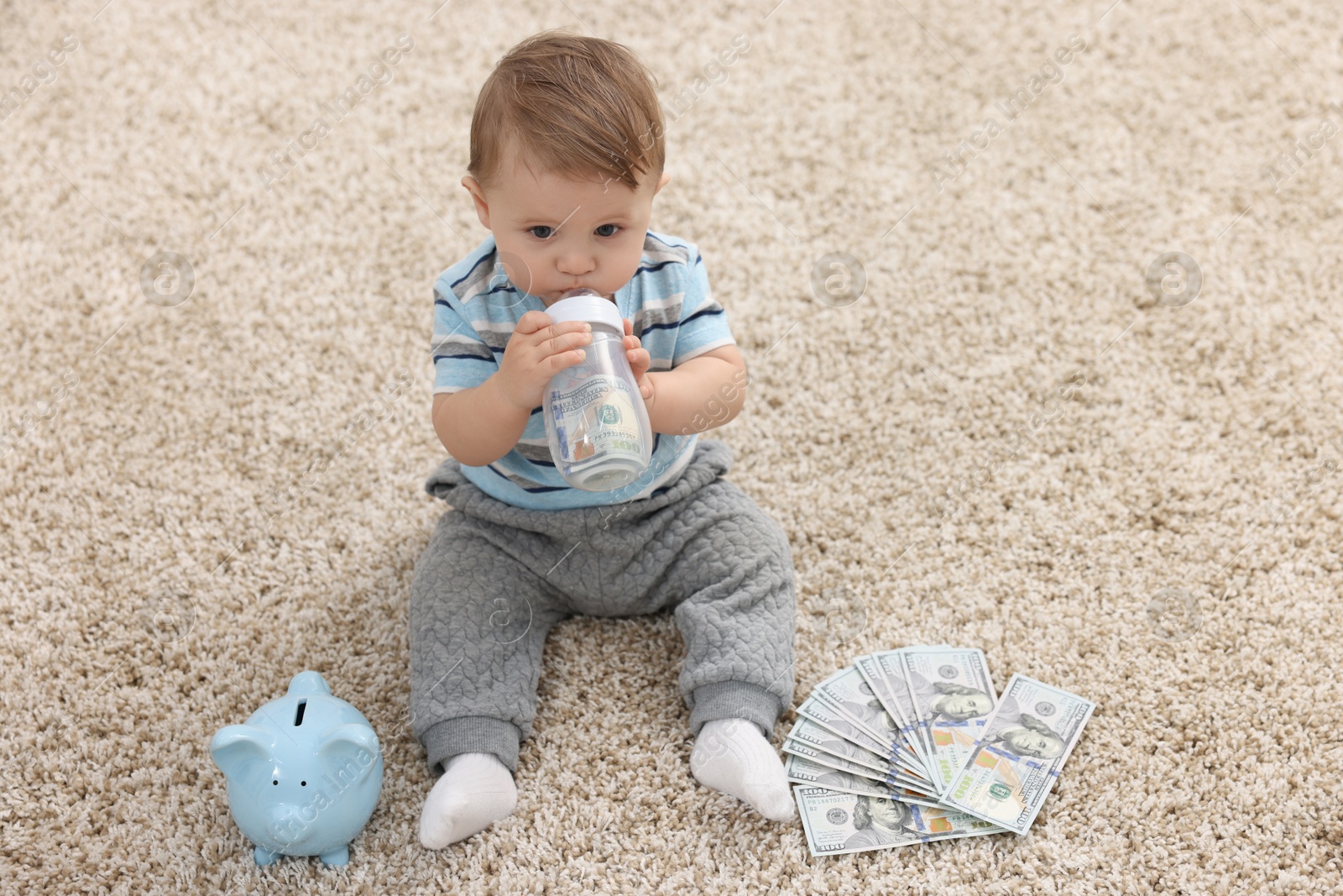 Photo of Little baby with bottle, money and piggybank on floor at home