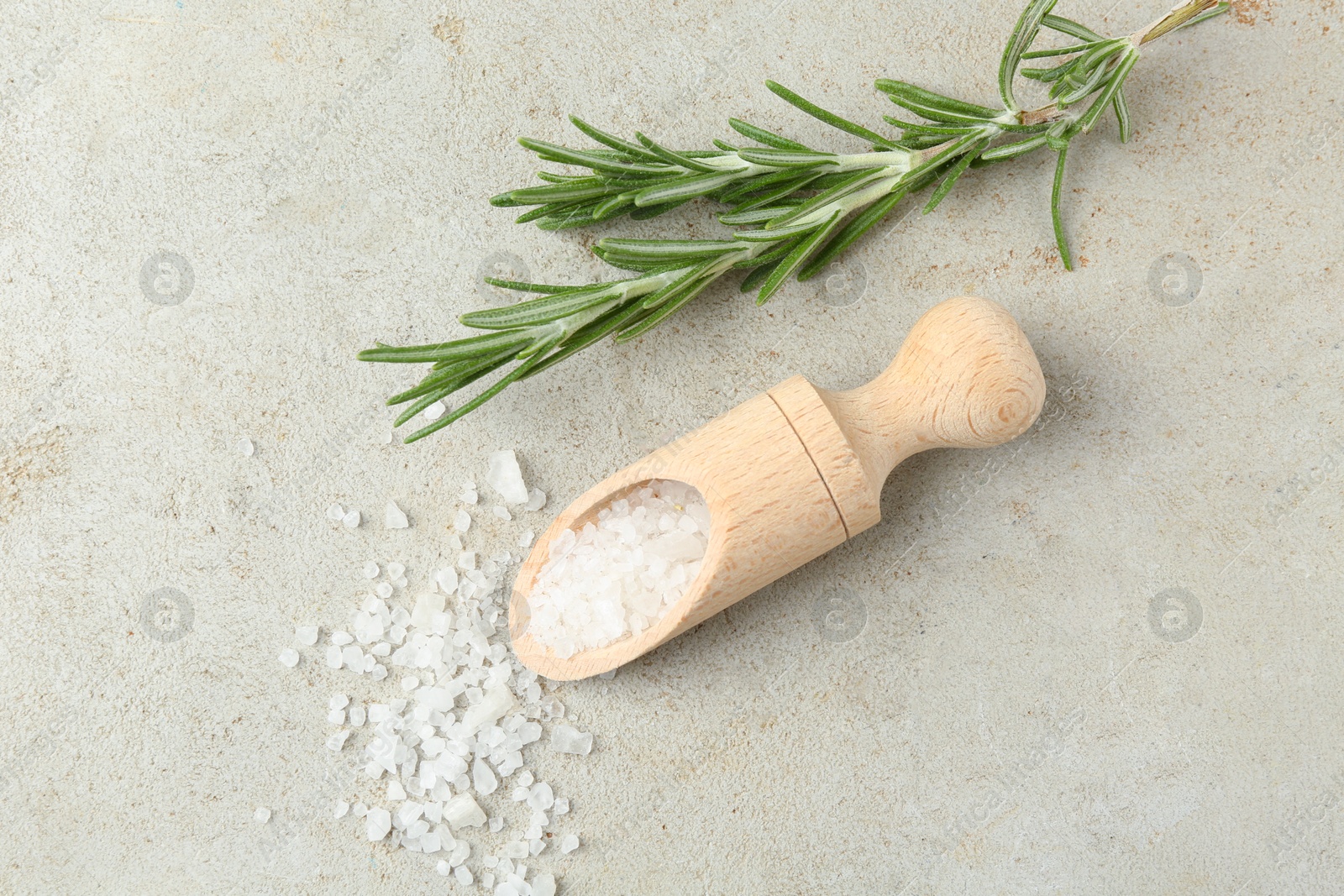 Photo of Sea salt in wooden scoop and rosemary on grey table, top view