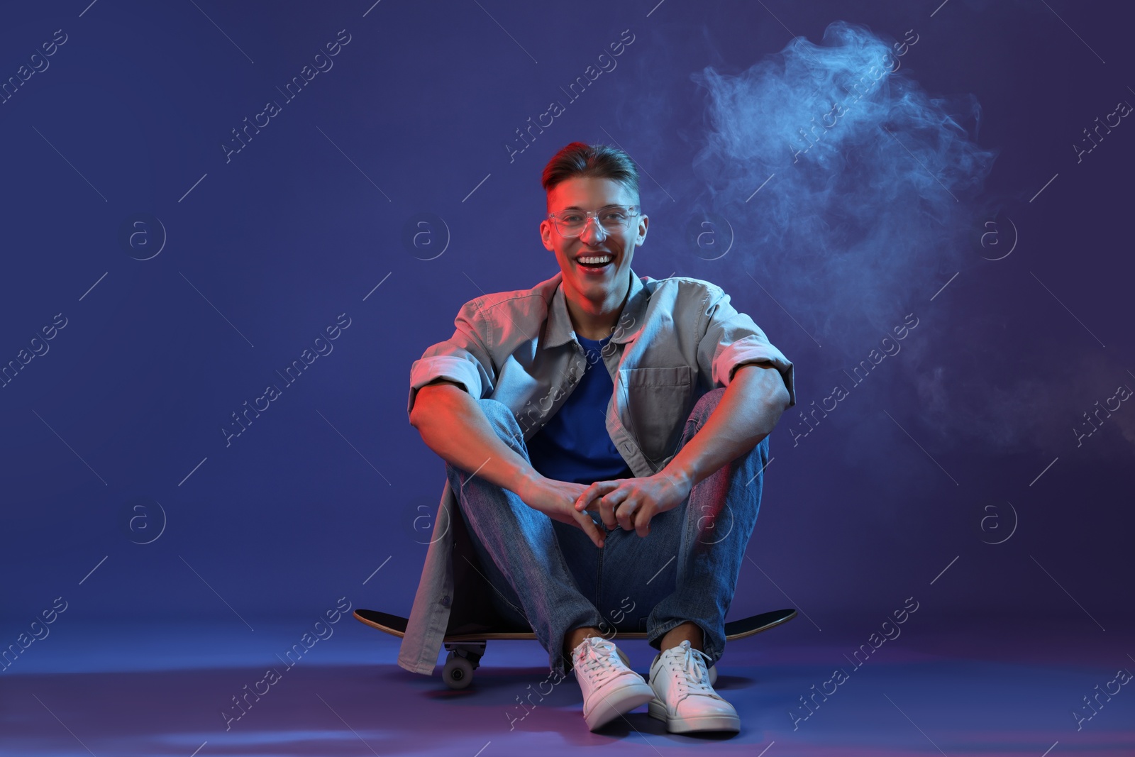 Photo of Happy man sitting on skateboard in smoke and color lights against blue background. Space for text