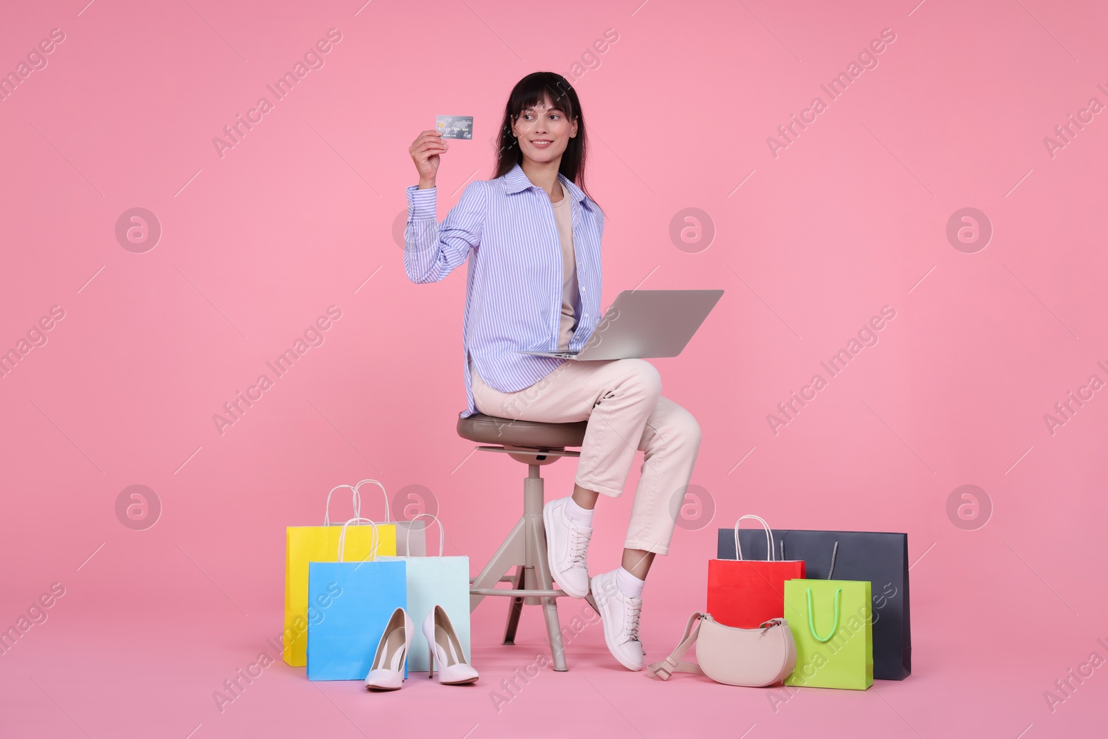 Photo of Internet shopping. Happy woman with credit card and laptop sitting on stool among colorful bags against pink background