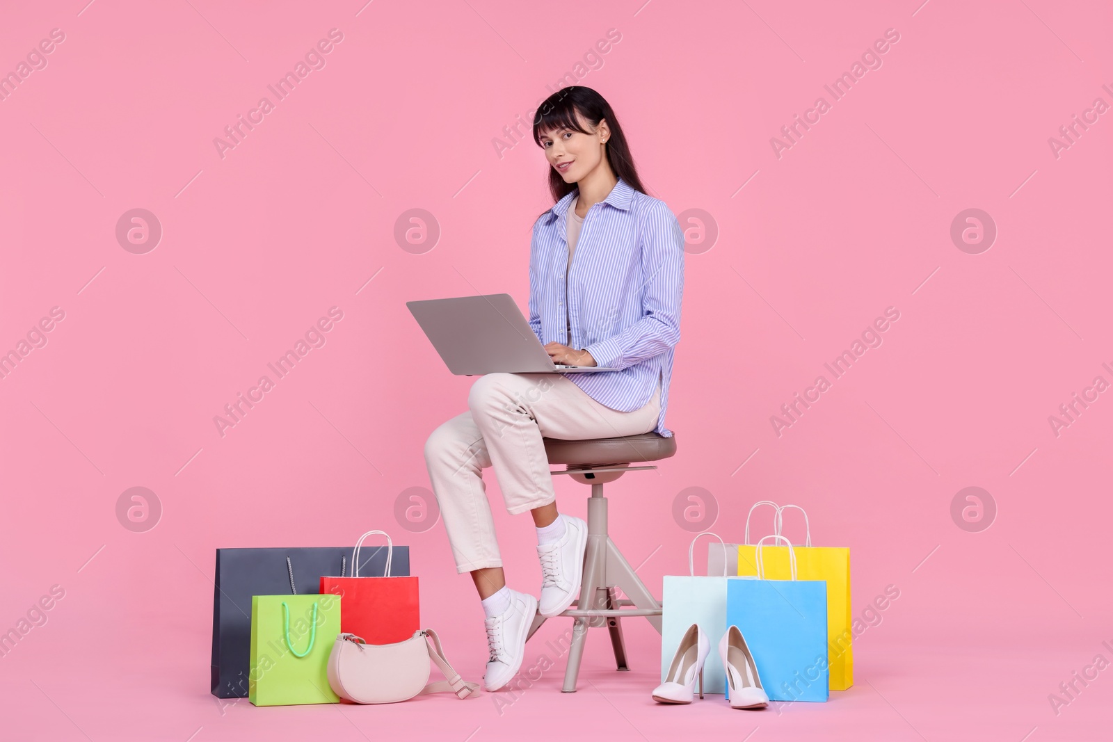 Photo of Internet shopping. Happy woman with laptop sitting on stool among colorful bags against pink background