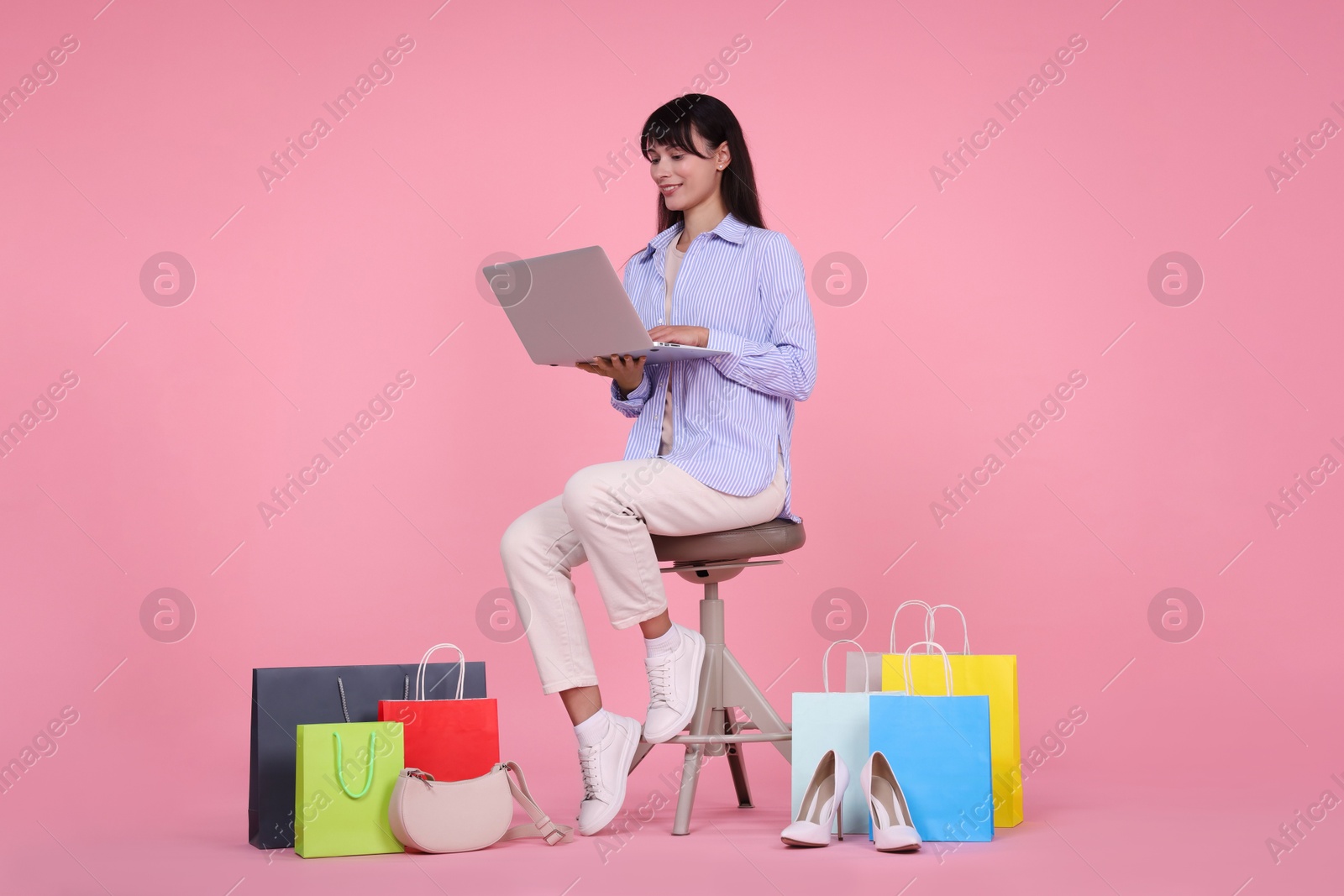 Photo of Internet shopping. Happy woman with laptop sitting on stool among colorful bags against pink background