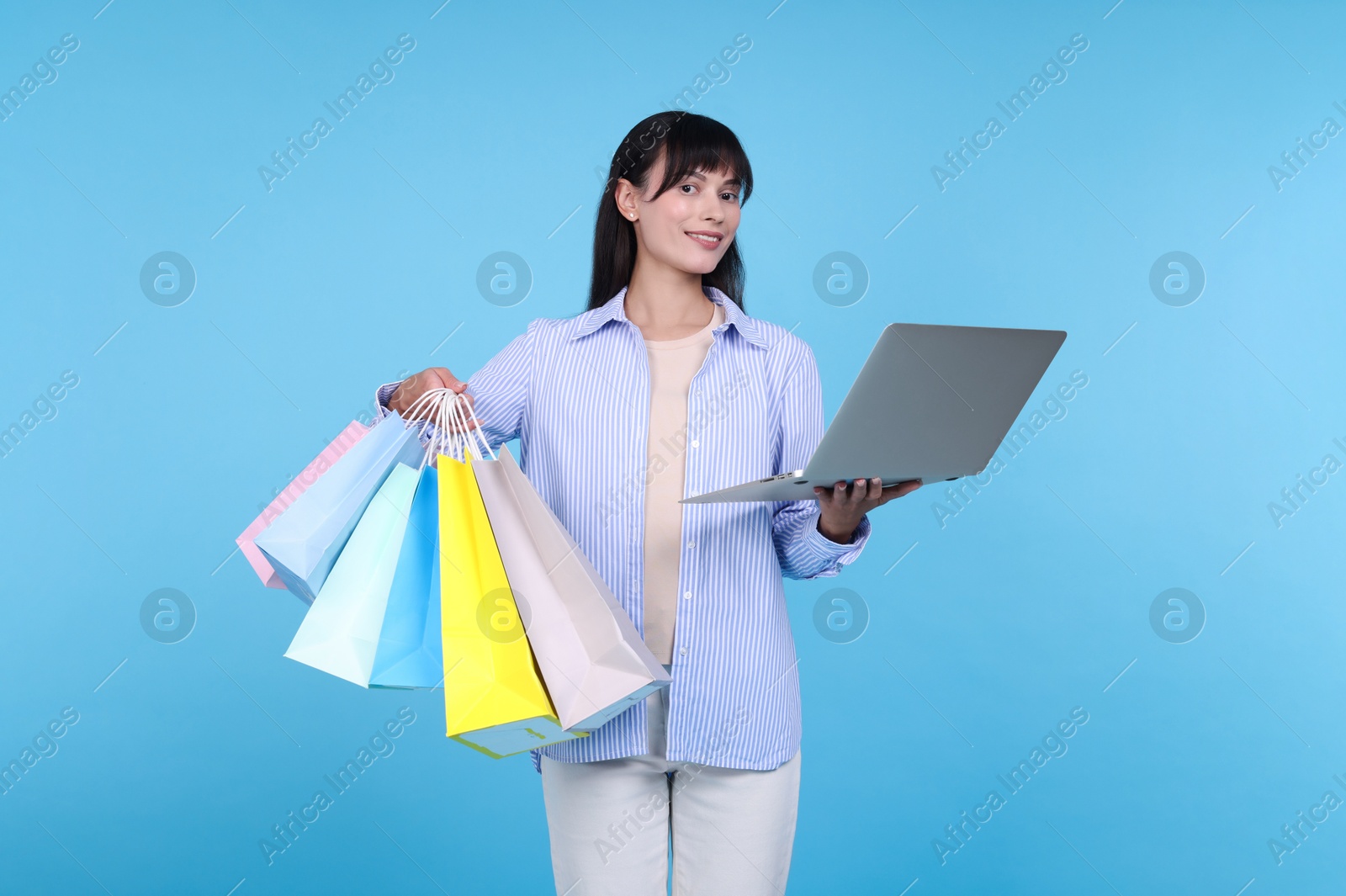 Photo of Internet shopping. Happy woman with laptop and colorful bags on light blue background