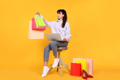 Photo of Internet shopping. Happy woman with laptop sitting on stool among colorful bags against orange background