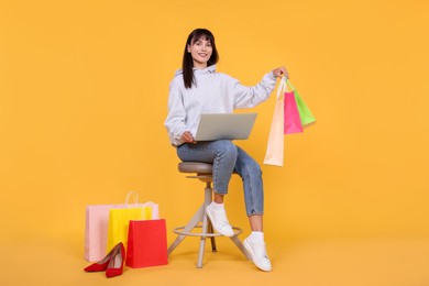 Photo of Internet shopping. Happy woman with laptop sitting on stool among colorful bags against orange background