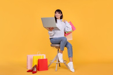 Photo of Internet shopping. Happy woman with laptop sitting on stool among colorful bags against orange background