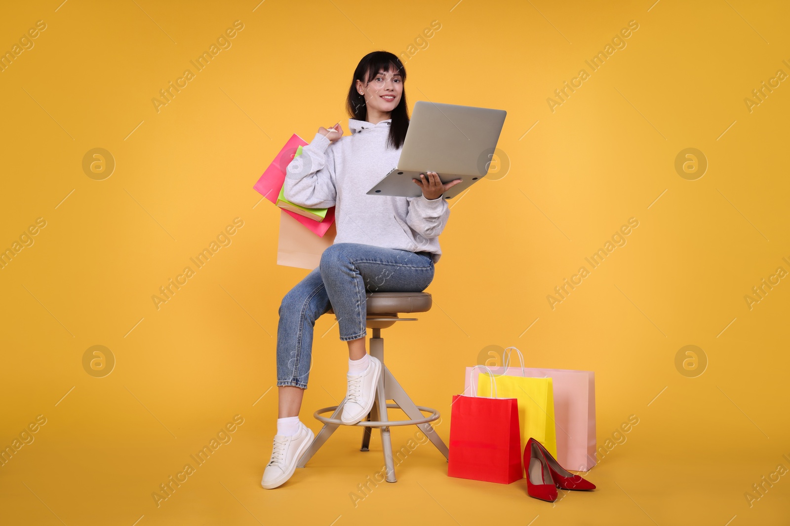 Photo of Internet shopping. Happy woman with laptop sitting on stool among colorful bags against orange background