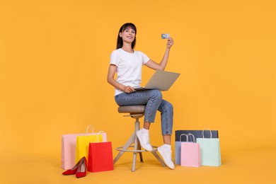 Photo of Internet shopping. Happy woman with credit card and laptop sitting on stool among colorful bags against orange background