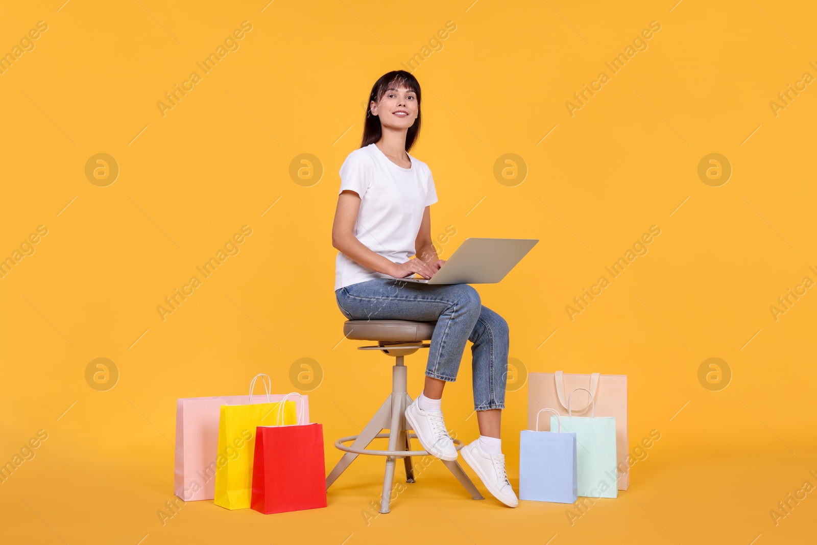 Photo of Internet shopping. Happy woman with laptop sitting on stool among colorful bags against orange background