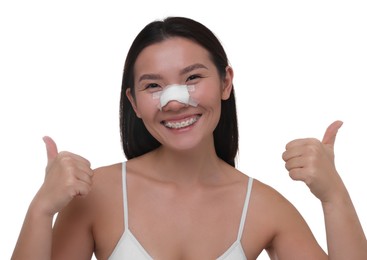 Photo of Woman with medical bandage on her nose after plastic surgery operation showing thumbs up against white background