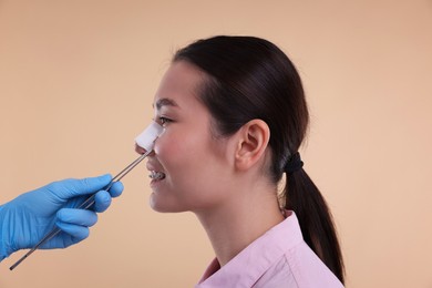 Photo of Doctor removing medical bandage from patient's nose after plastic surgery operation on beige background, closeup