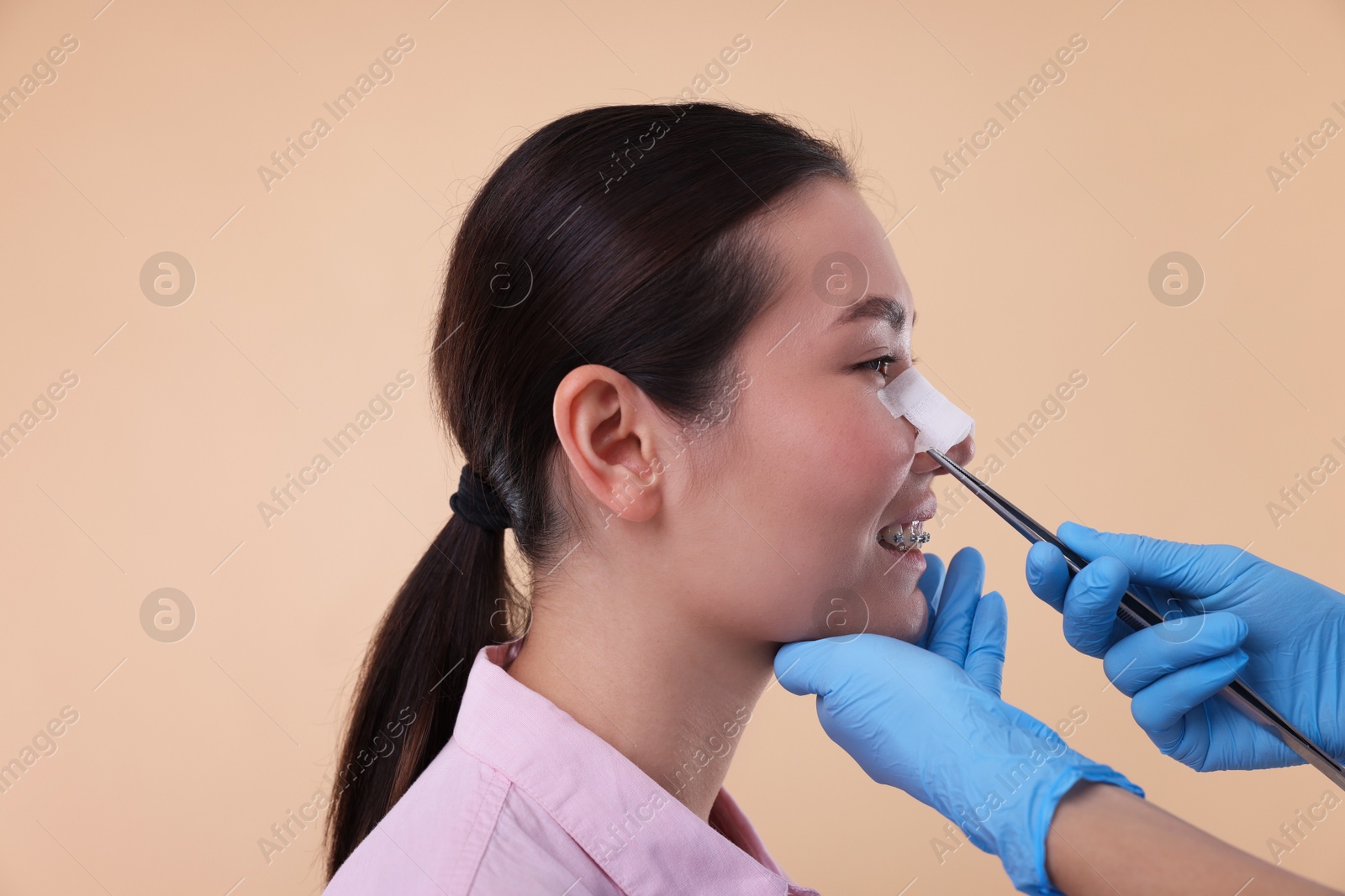 Photo of Doctor removing medical bandage from patient's nose after plastic surgery operation on beige background, closeup