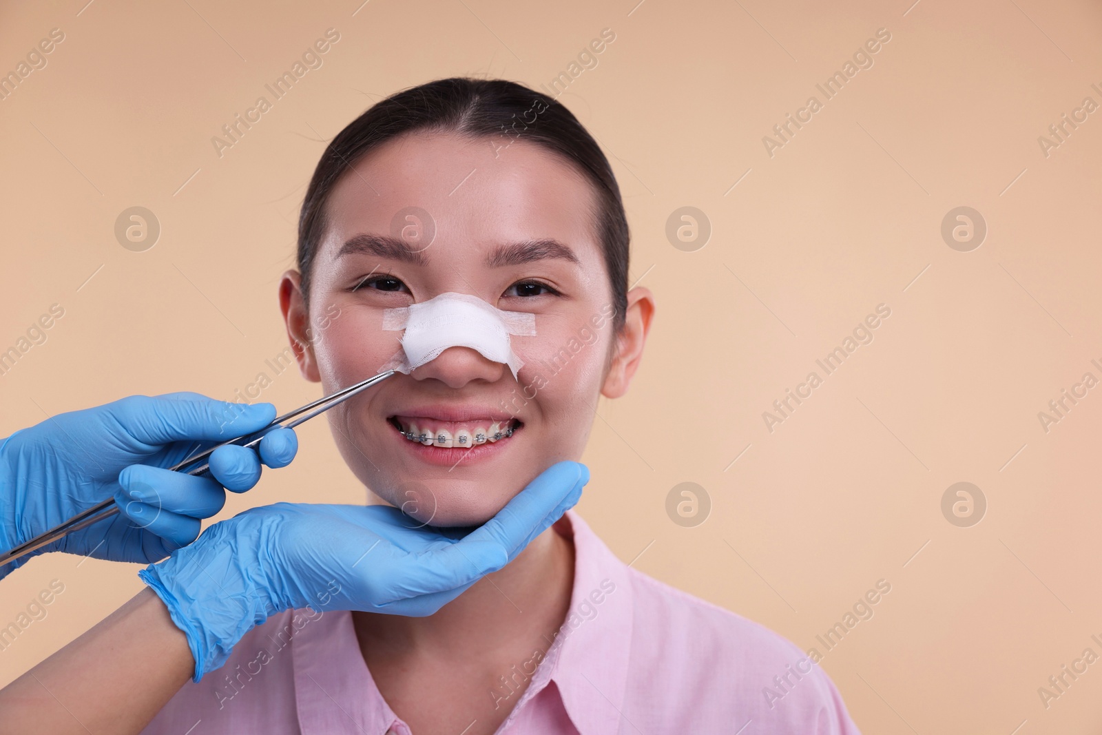 Photo of Doctor removing medical bandage from patient's nose after plastic surgery operation on beige background, closeup. Space for text