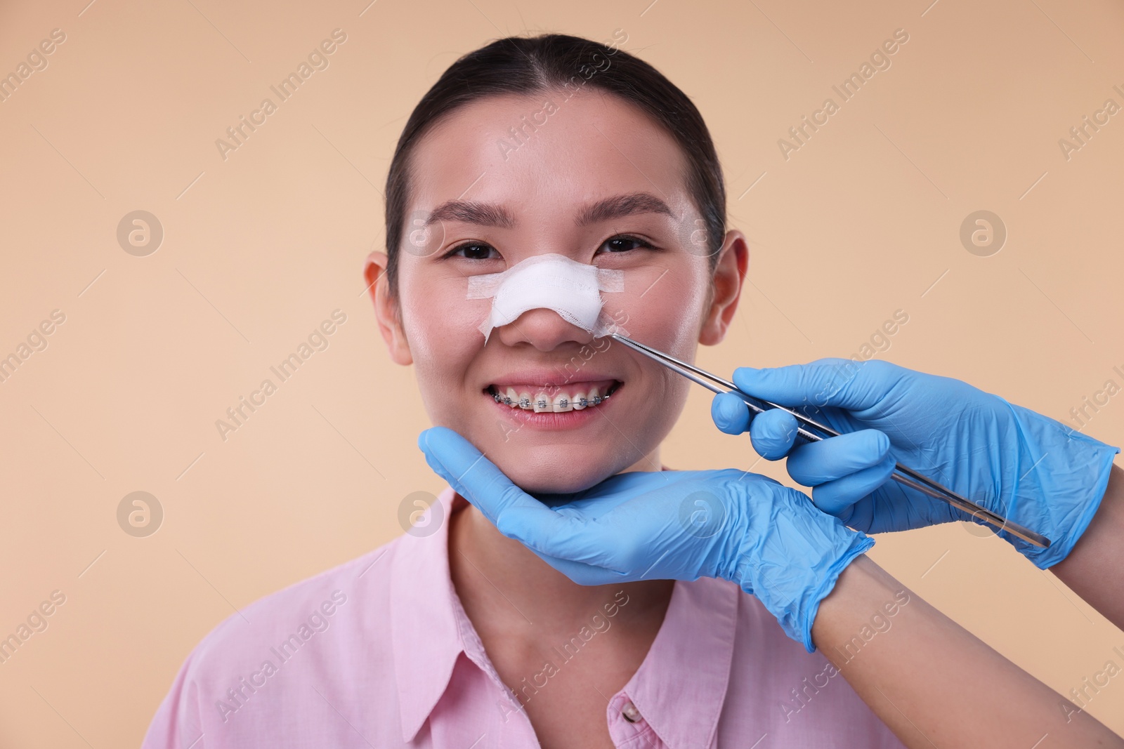 Photo of Doctor removing medical bandage from patient's nose after plastic surgery operation on beige background, closeup