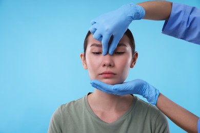 Photo of Doctor checking patient's nose before plastic surgery operation on light blue background, closeup