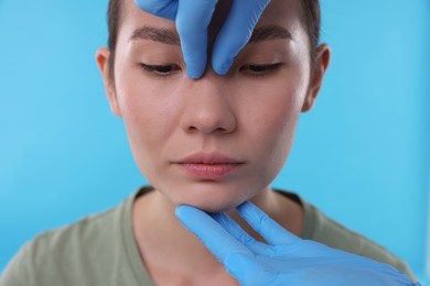 Photo of Doctor checking patient's nose before plastic surgery operation on light blue background, closeup