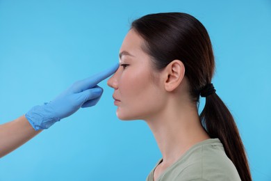 Photo of Doctor checking patient's nose before plastic surgery operation on light blue background, closeup