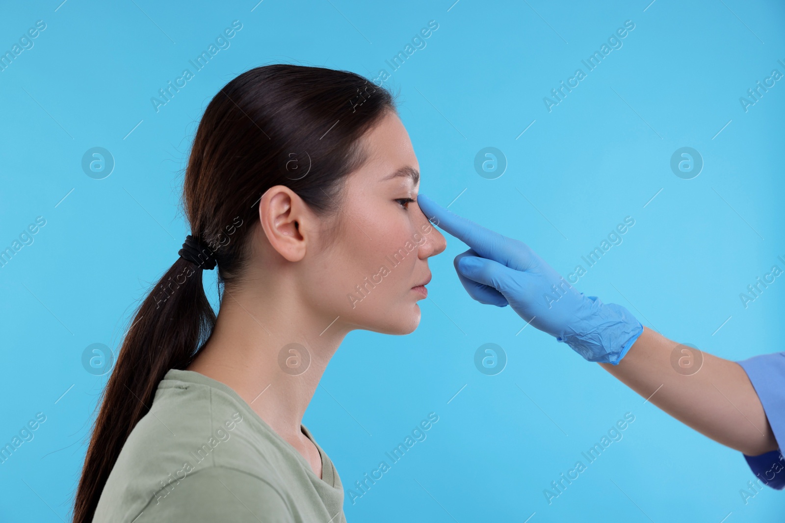 Photo of Doctor checking patient's nose before plastic surgery operation on light blue background, closeup