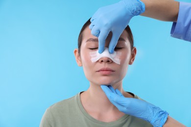 Photo of Doctor checking patient's nose after plastic surgery operation on light blue background, closeup