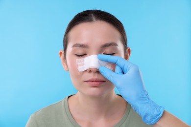 Photo of Doctor checking patient's nose after plastic surgery operation on light blue background, closeup