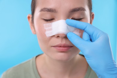 Photo of Doctor checking patient's nose after plastic surgery operation on light blue background, closeup