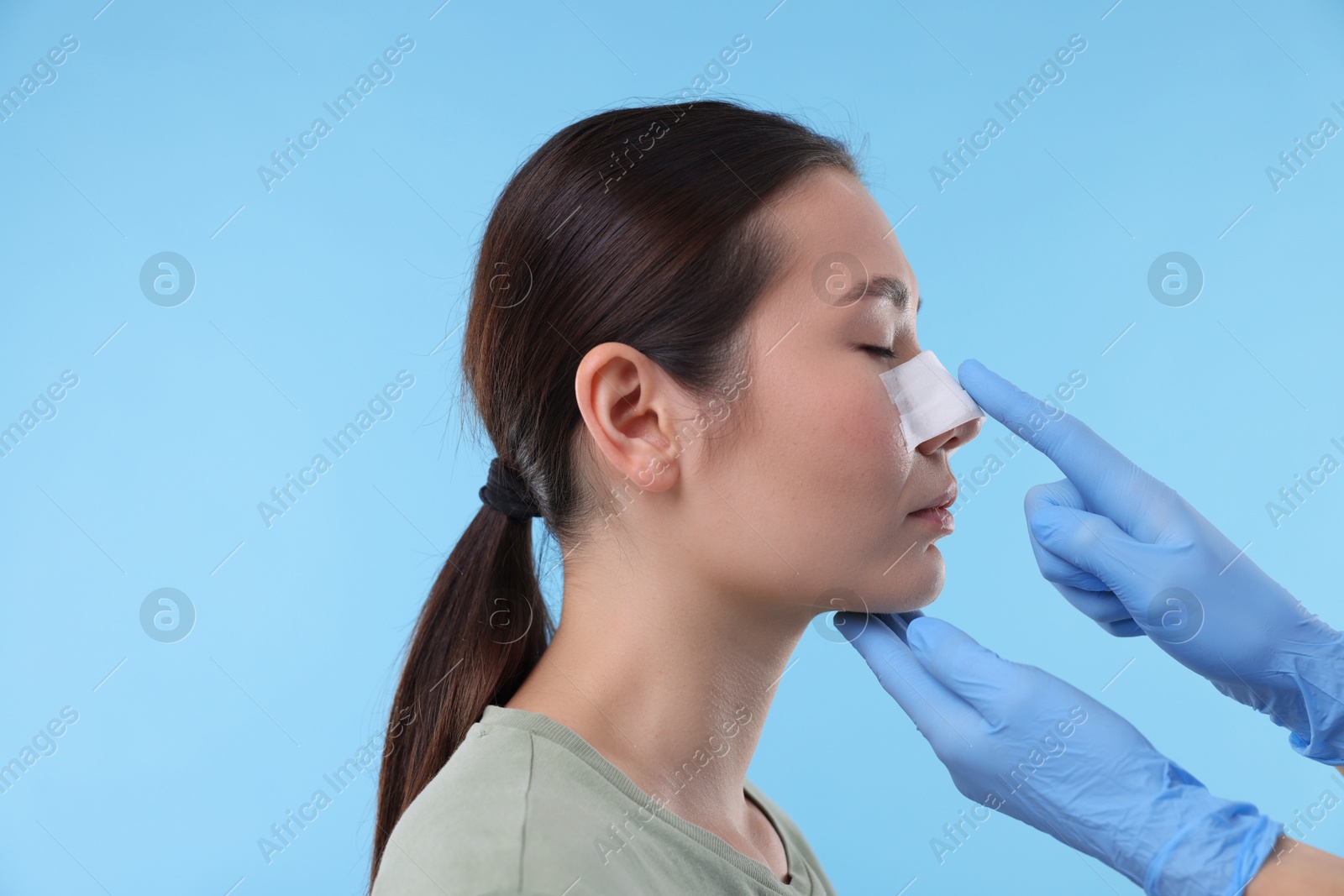 Photo of Doctor checking patient's nose after plastic surgery operation on light blue background, closeup