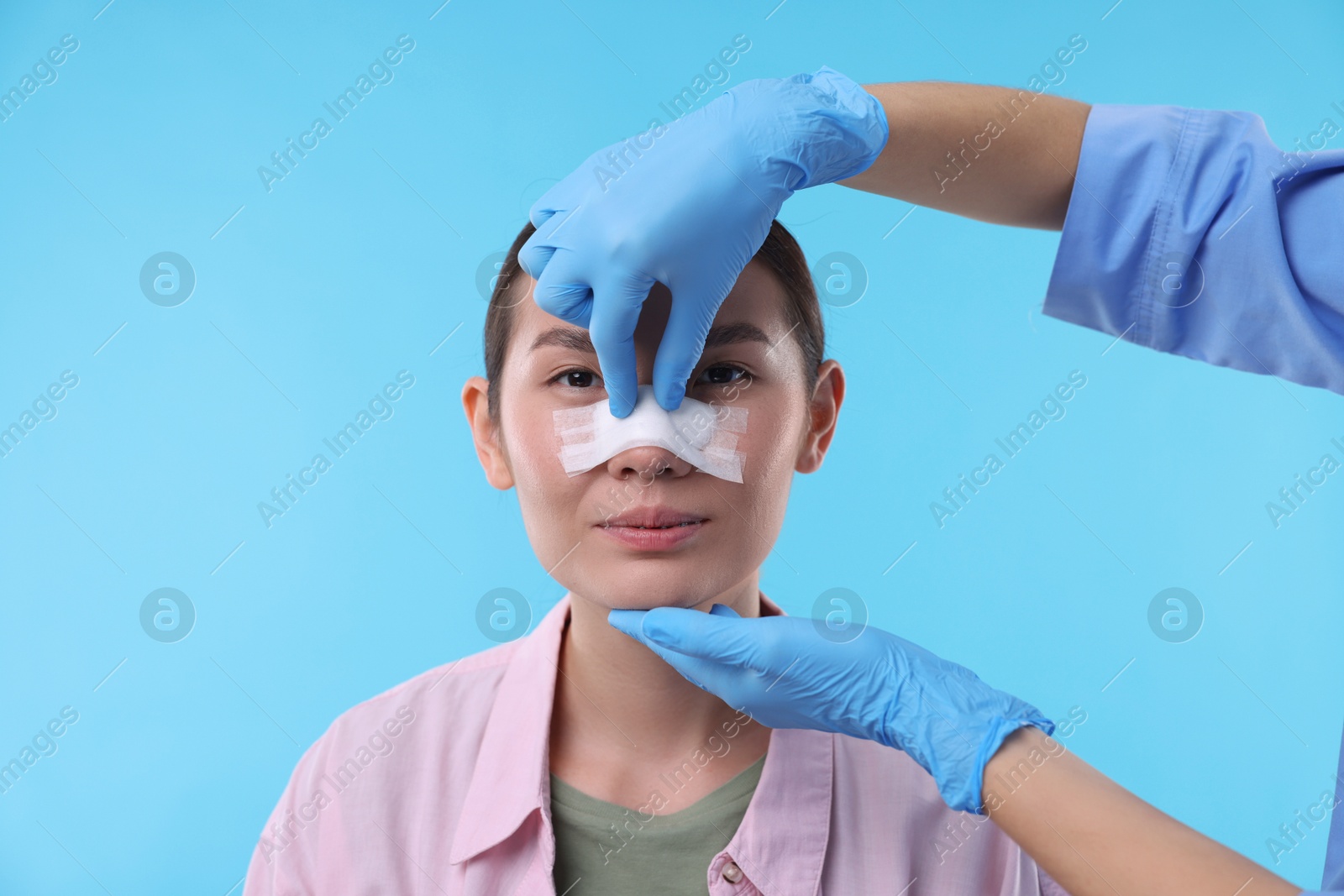 Photo of Doctor checking patient's nose after plastic surgery operation on light blue background, closeup