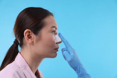 Photo of Doctor checking patient's nose after plastic surgery operation on light blue background, closeup