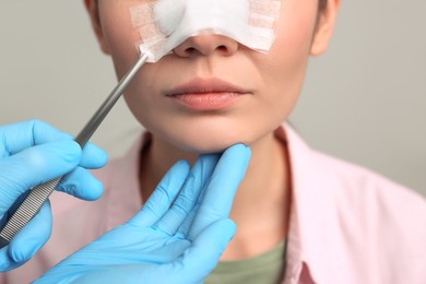 Doctor removing medical bandage from patient's nose after plastic surgery operation on light grey background, closeup