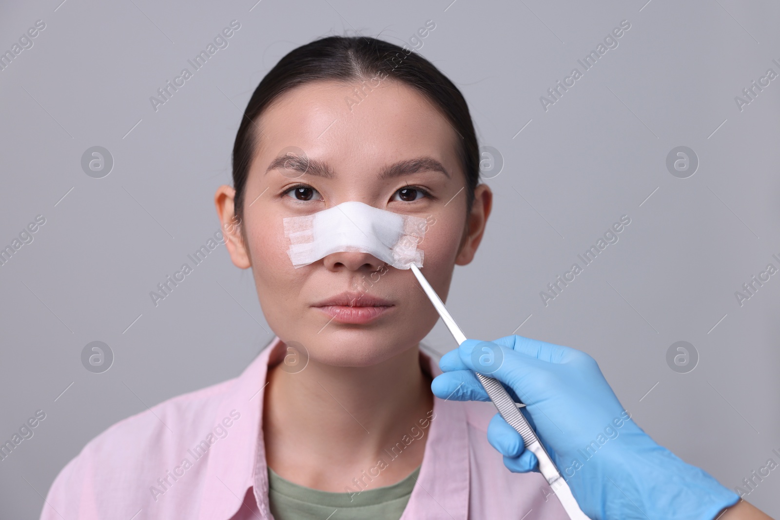 Photo of Doctor removing medical bandage from patient's nose after plastic surgery operation on light grey background, closeup