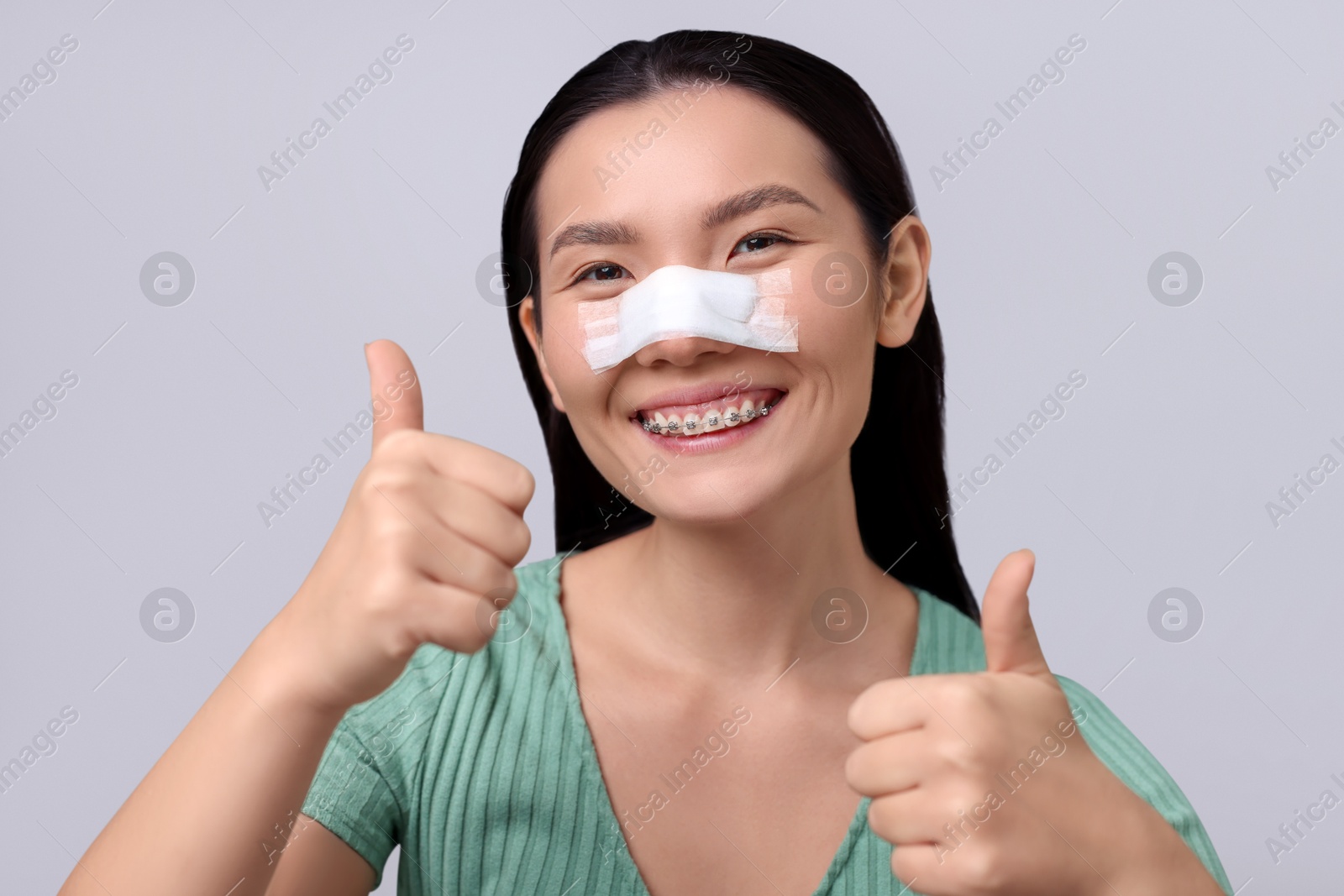 Photo of Woman with medical bandage on her nose after plastic surgery operation showing thumbs up against light grey background