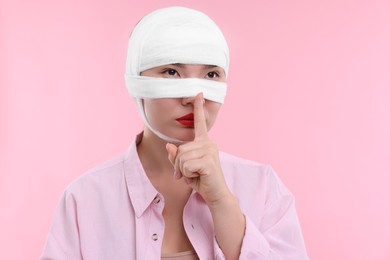 Woman with nose wrapped in medical bandage after plastic surgery operation making silent gesture on pink background