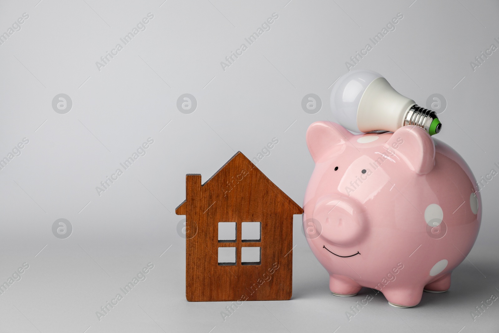 Photo of Piggy bank, house model and light bulb on grey background, space for text. Energy saving concept