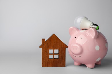 Piggy bank, house model and light bulb on grey background, space for text. Energy saving concept