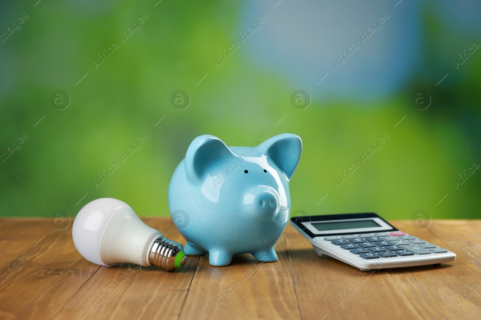 Photo of Piggy bank, calculator and light bulb on wooden table outdoors. Energy saving concept