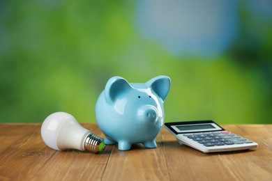 Photo of Piggy bank, calculator and light bulb on wooden table outdoors. Energy saving concept