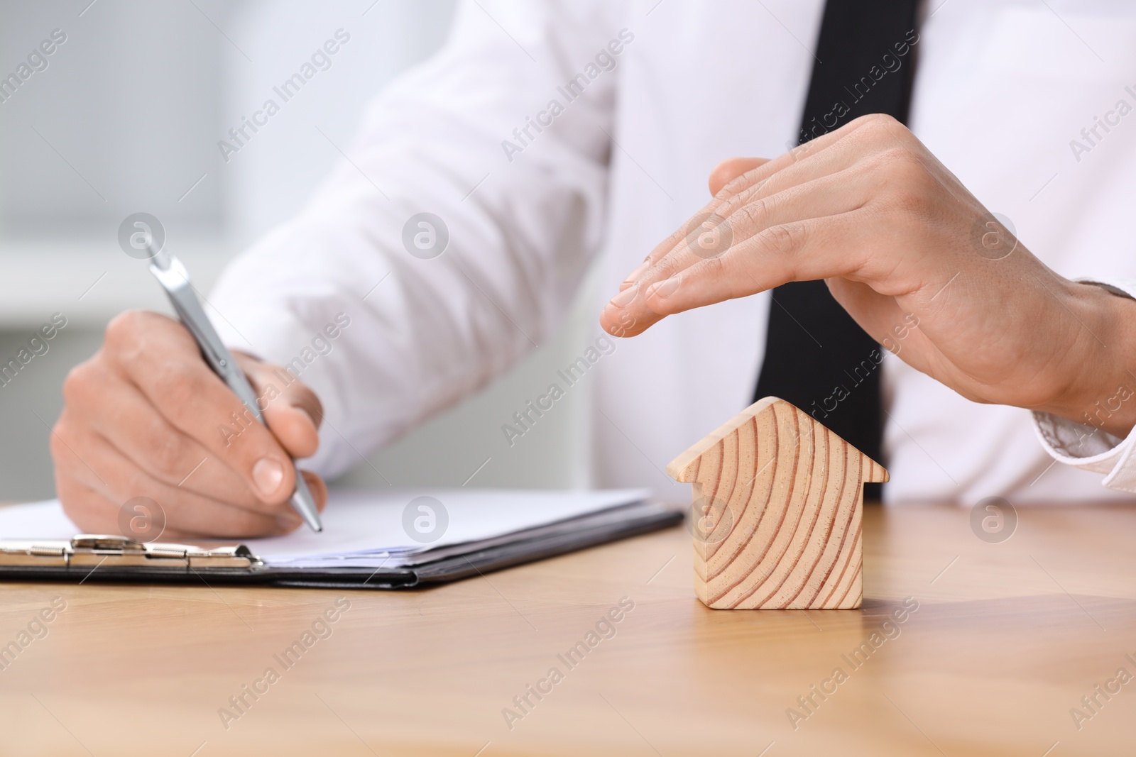 Photo of Property insurance. Real estate agent protecting house figure while working at wooden table, closeup