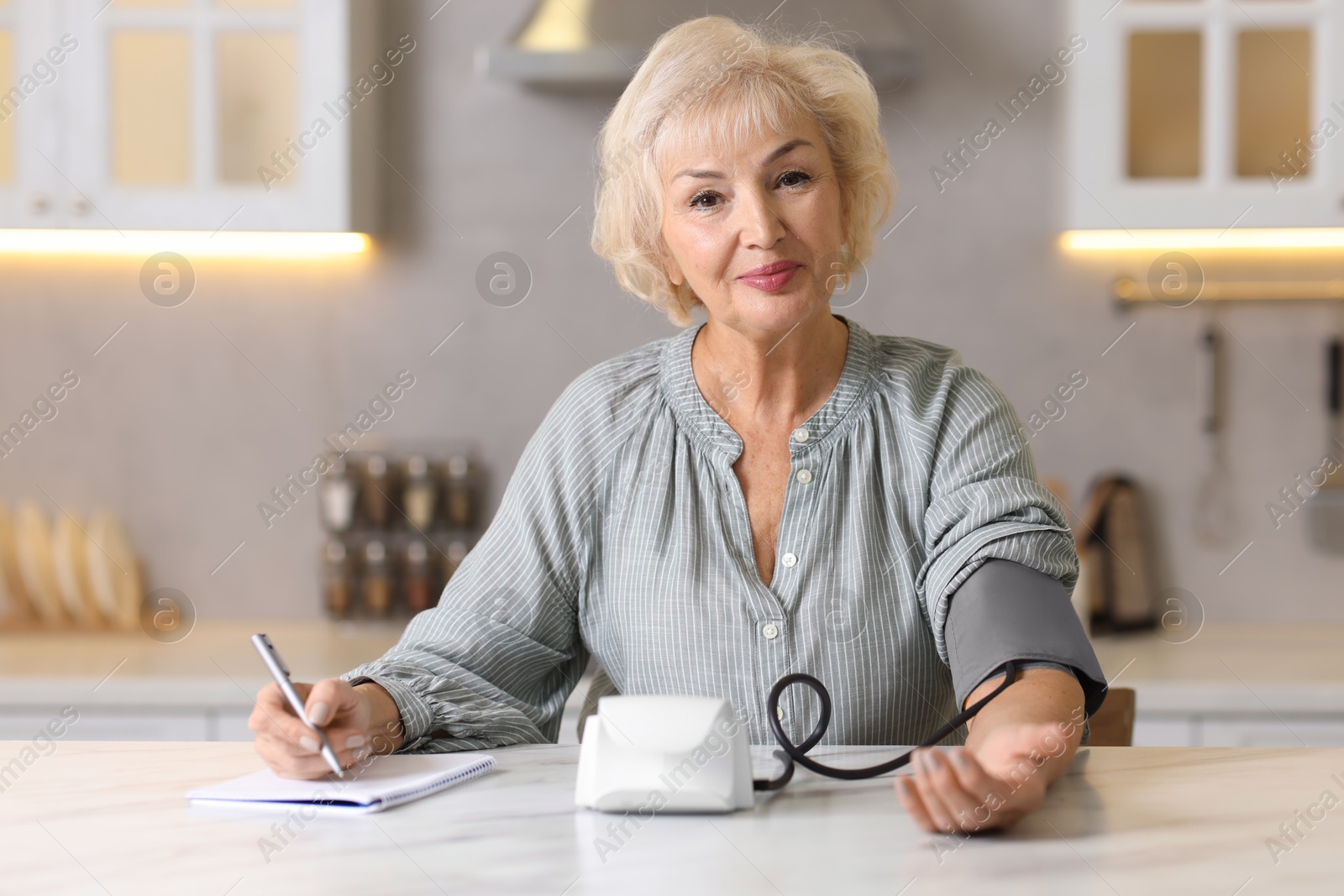 Photo of Senior woman writing results of blood pressure measurement in kitchen at home. Space for text