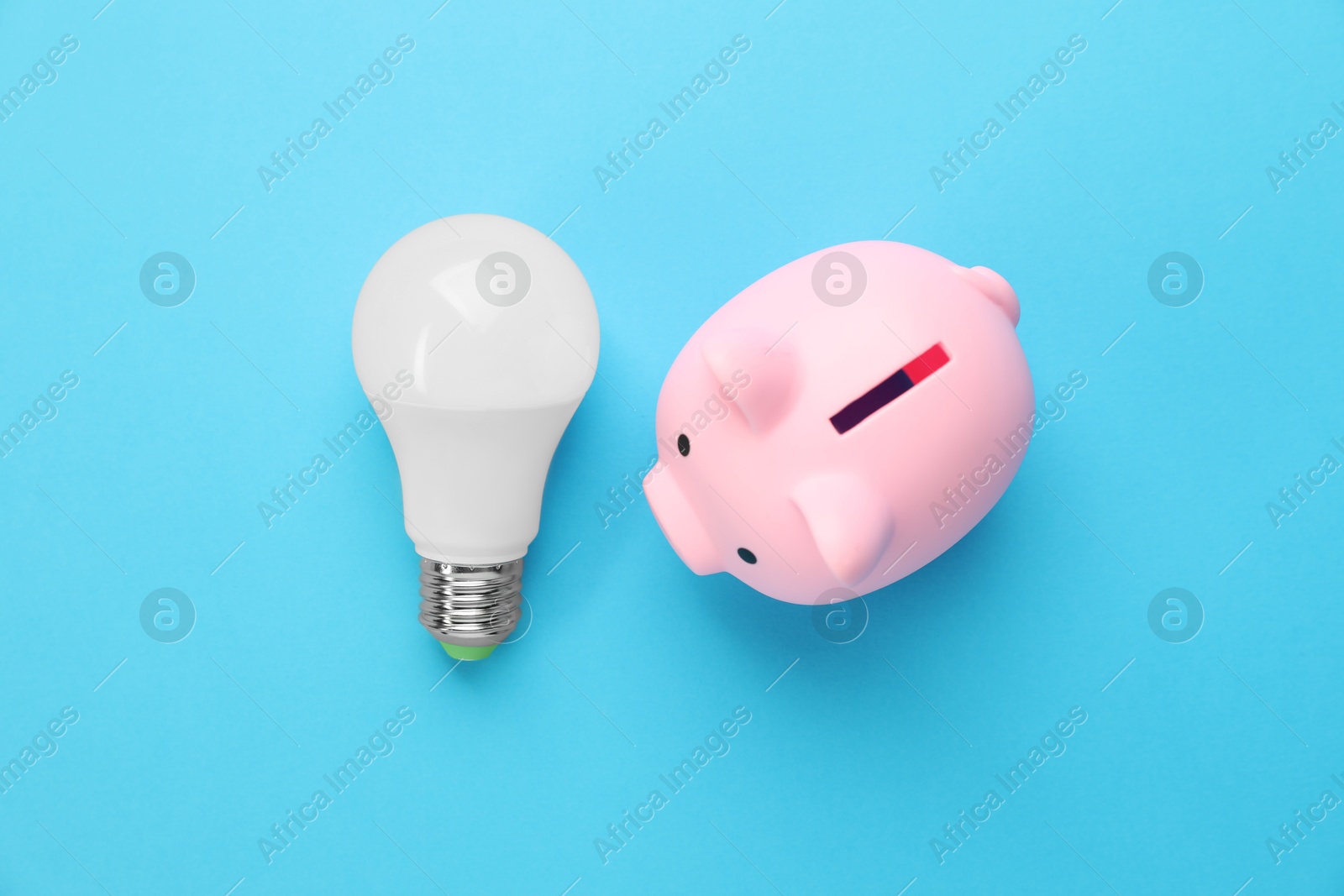 Photo of Piggy bank and lightbulb on light blue background, flat lay. Energy saving concept